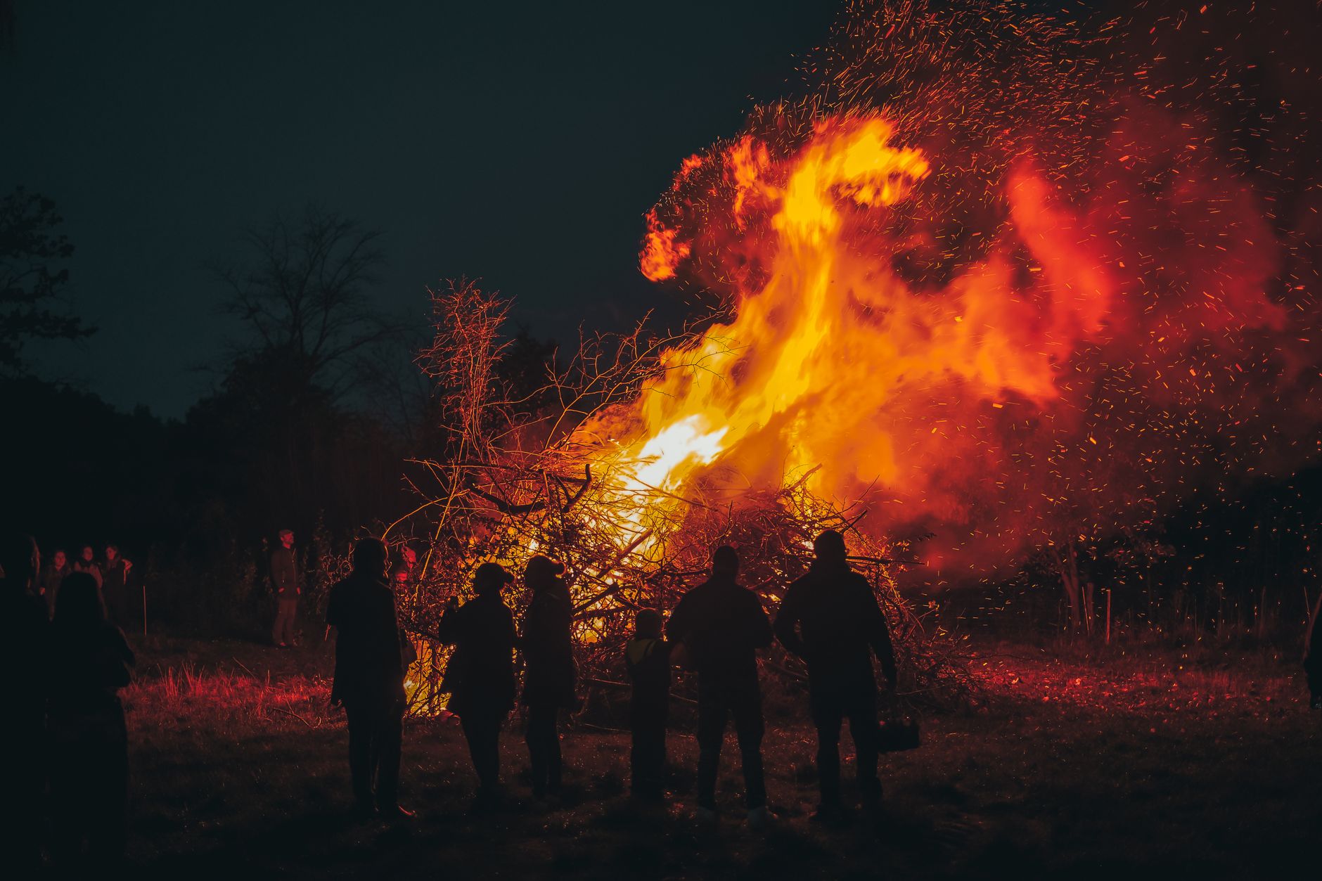 A tall bonfire burning with bright flames as people gather around it on a dark evening