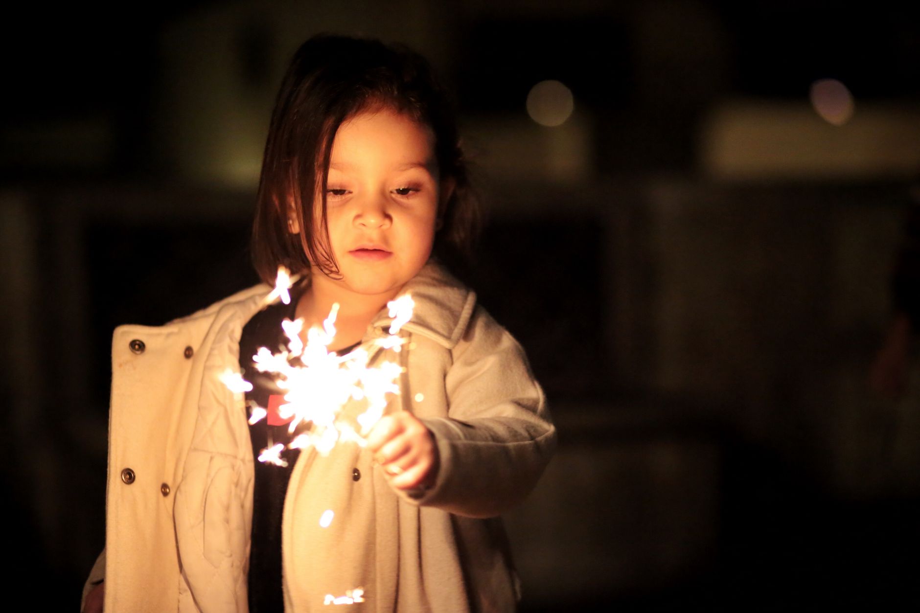 A child in a warm winter coat holding a lit sparkler at arm's length outdoors at night