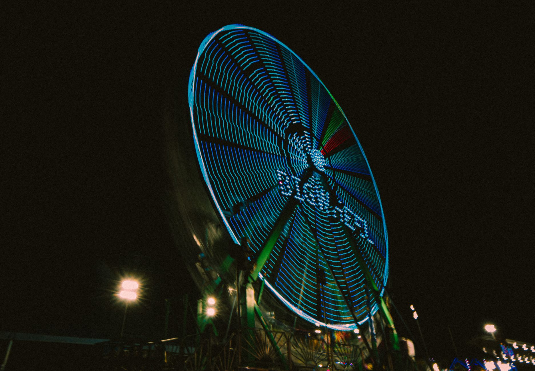 A brightly lit observation wheel and funfair rides against a dark winter sky in a city park